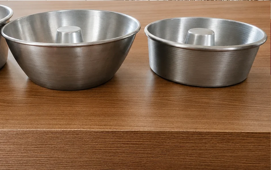 Três formas de bolo de alumínio com furo central, em tamanhos diferentes, alinhadas sobre uma mesa de madeira em uma cozinha iluminada.