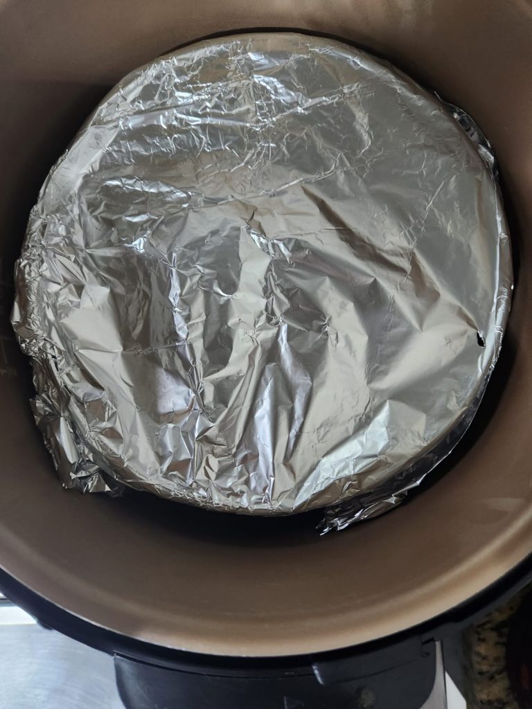 Forma de pudim de alumínio tampada com papel alumínio dentro do bowl da panela de pressão elétrica com uma xícara de água no fundo
